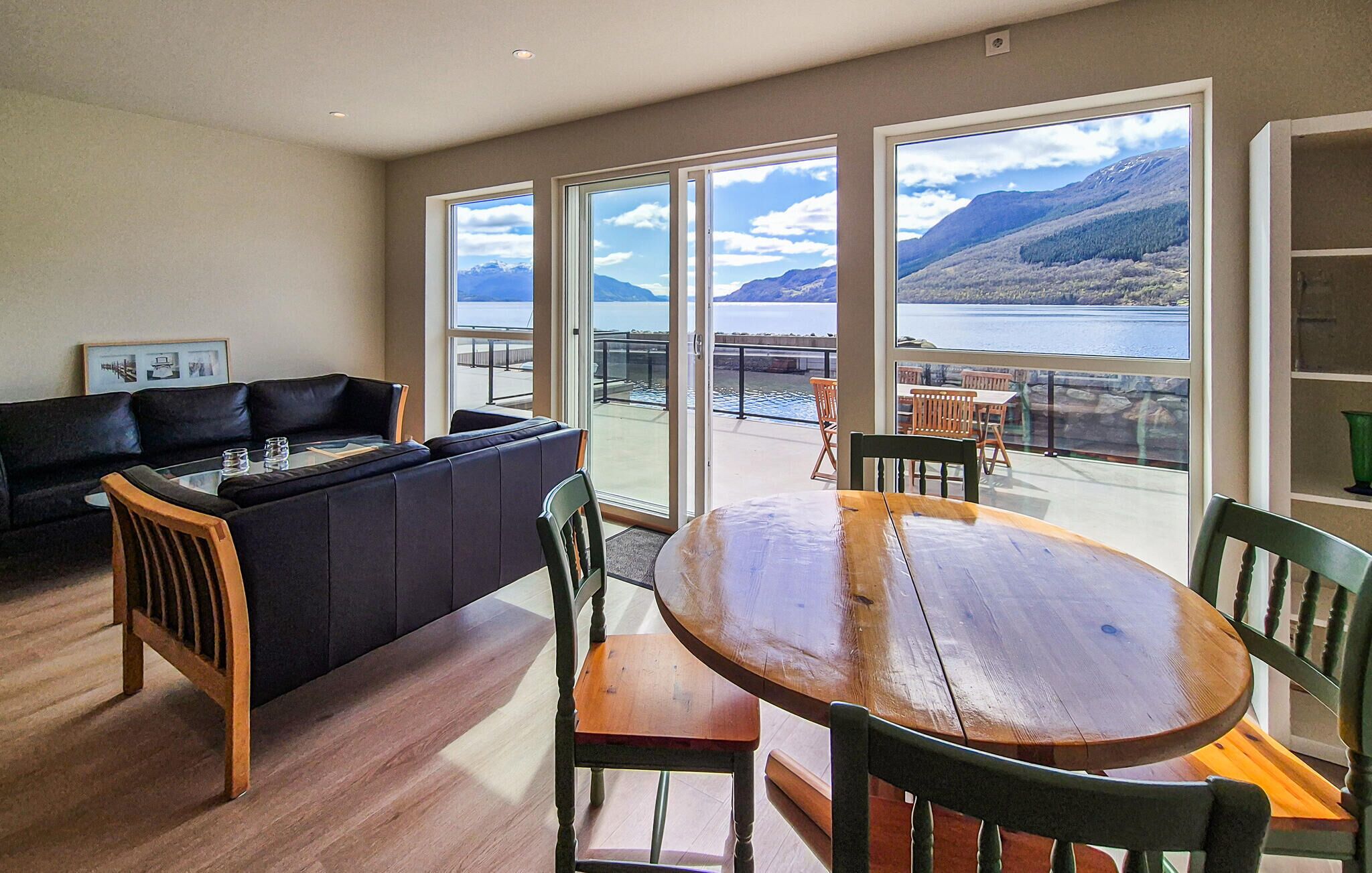 Una sala de estar con sofá, una mesa de comedor y sillas, con ventanas amplias que ofrecen vistas a un lago y montañas.