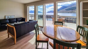 A living room with a sofa, a dining table, and chairs, with large windows offering a view of a lake and mountains.