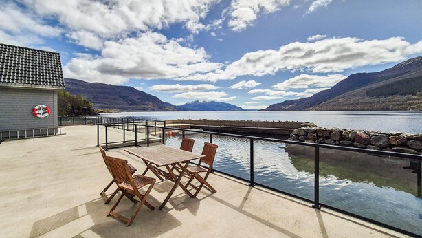 A deck with a table and chairs overlooking a lake, a lifebuoy on a wall, and mountains in the background.
