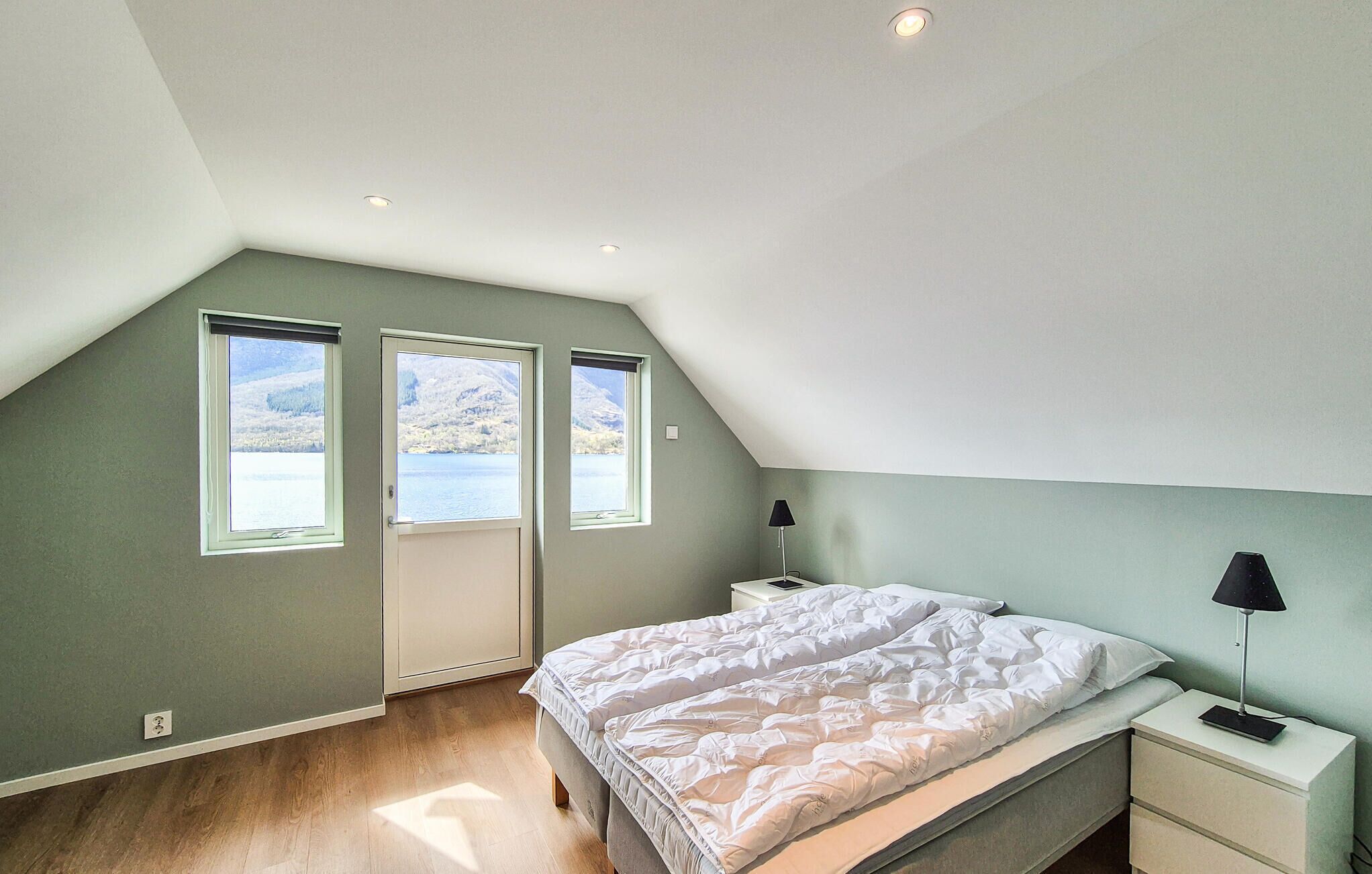 Un dormitorio con techo inclinado, cama, mesita de noche, lámpara y ventana con vista a un lago y montañas.