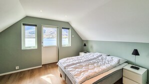A bedroom with a sloped ceiling, a bed, a nightstand, a lamp, and a window with a view of a lake and mountains.