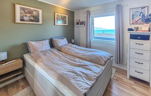 En dobbeltseng med hvide sengetøj, et natbord med lampe, en kommode med skuffer og et vindue med havudsigt.
