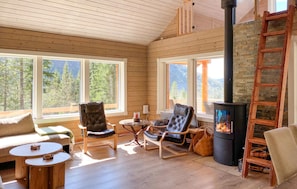 A cozy cabin interior with wooden walls, large windows, a fireplace, and comfortable seating.
