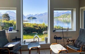 A room with large windows offering a view of a lake and mountains. There are two chairs and a small table. - Stunning home in ålesund with house sea view (Ålesund)