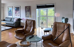 A modern living room with leather armchairs, a glass coffee table, a grey sofa, and a large window with a view of greenery. - Stunning home in ålesund with house sea view (Ålesund)