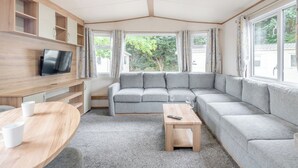 Ein modernes Wohnzimmer mit einem grauen Ecksofa, einem an einer Holzwand montierten Fernseher, einem kleinen Holztisch und einem Fenster mit Vorhängen.