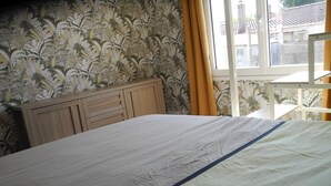 A bed with a wooden headboard, a window with yellow curtains, and a view of buildings outside. - House 5 mn walk from the center of aiguillon (L'Aiguillon-la-Presqu'île)