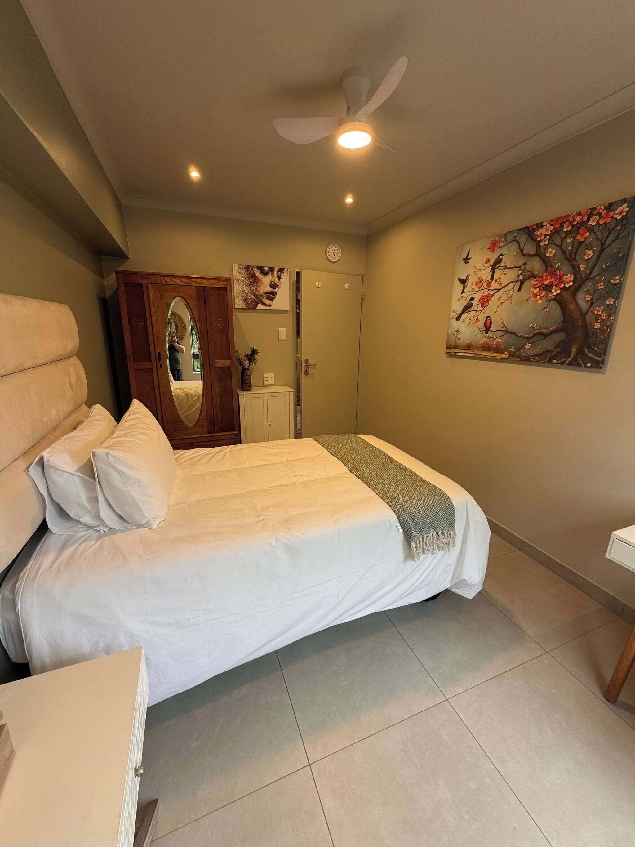 A bedroom with a bed, a wooden wardrobe, a mirror, a ceiling fan, a clock, and a painting of a tree with birds.