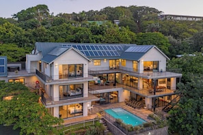 A modern multi-story house with solar panels, a pool, and a lush garden.