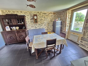 A dining area with a table set for two, a wooden cabinet with glass doors, a stone wall, and a window with a view. - 734 – Seaside holiday home in Primelin, Brittany (Primelin)