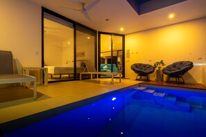 A modern indoor pool area with a glass wall, a ceiling fan, and a seating area with chairs.