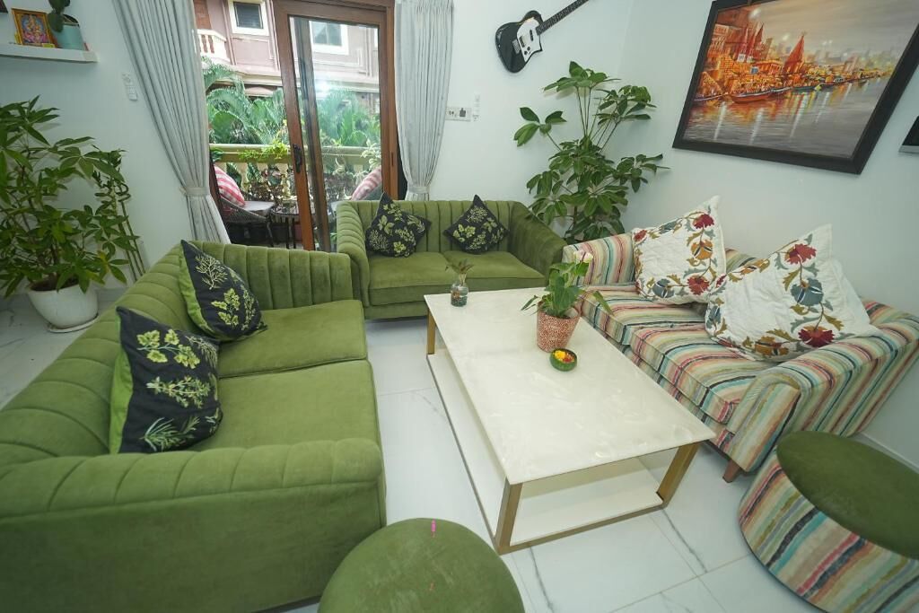 A living room with a green sofa set, a coffee table, and a striped sofa. There are potted plants and a guitar on the wall.