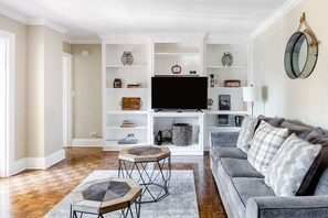 Ein Wohnzimmer mit grauem Sofa, einem Couchtisch, einem Fernseher auf einem weißen Schrank und Regalen mit diversen Gegenständen.