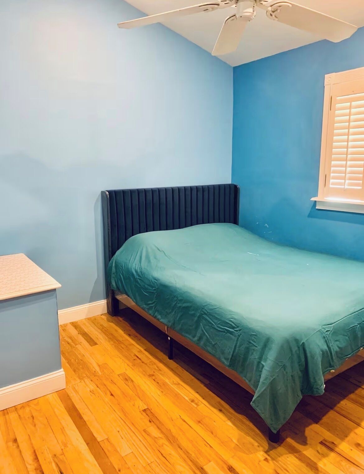 Une chambre à coucher comprenant un lit, un radiateur, un ventilateur de plafond, une fenêtre avec des stores et un plancher en bois.
