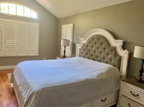A bedroom with a large bed, a nightstand, a lamp, and a window with shutters. - Spacious Luxe 5-bedroom house with beautiful resort backyard (Dix Hills)
