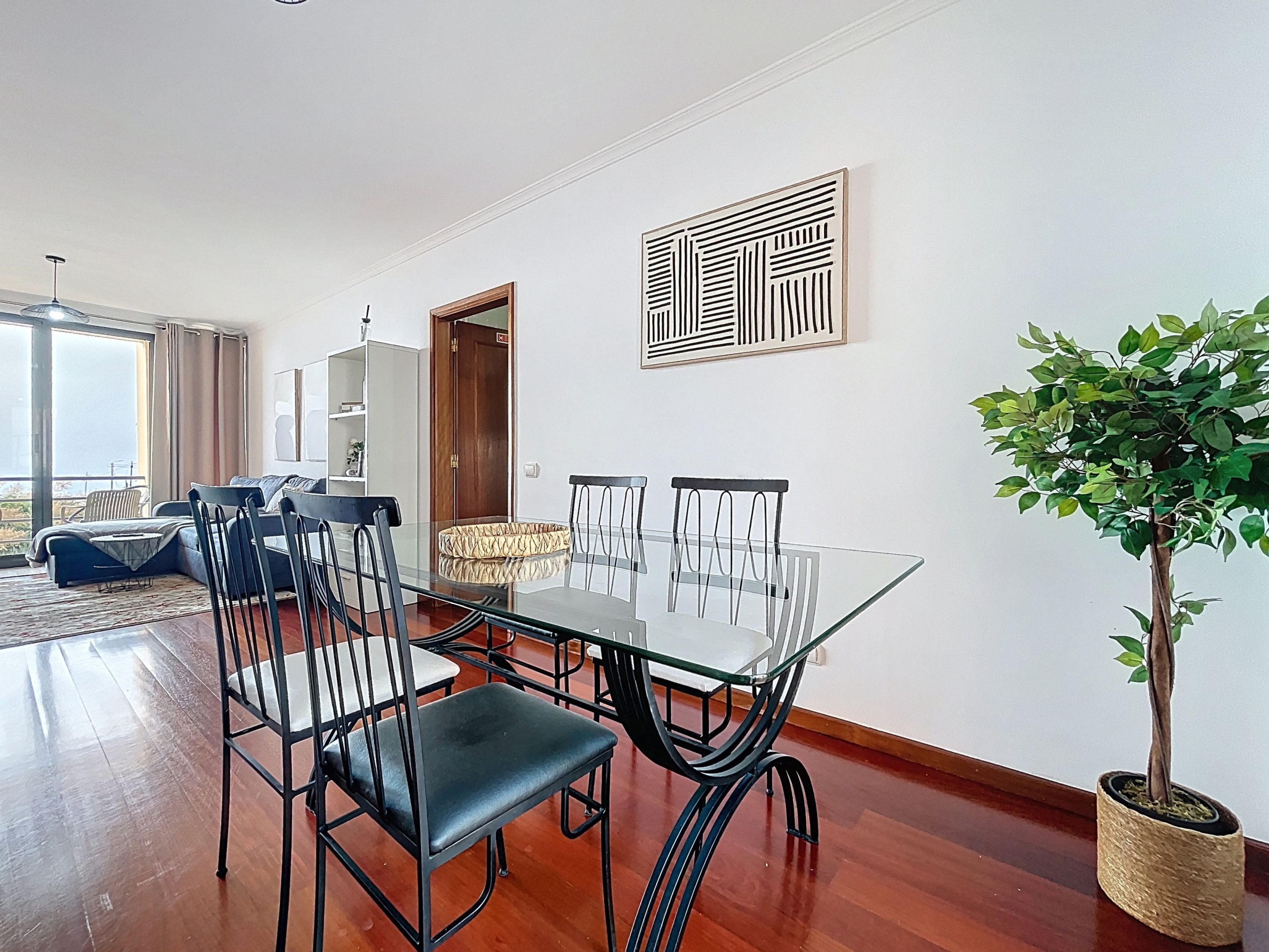 A dining area with a glass table and chairs, a potted plant, and a view of the balcony.