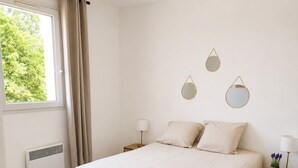 Une chambre à coucher comprenant un lit, des tables de chevet, une fenêtre avec des rideaux, un radiateur et des miroirs fixés au mur.
