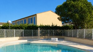 Une piscine aux eaux claires, avec un escalier en métal, entourée d’une clôture et bordée par un bâtiment en arrière-plan.