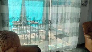 Une pièce avec un fauteuil en cuir marron, un plancher en bois, une porte en verre donnant sur un espace extérieur équipé d’une table et de chaises, et une vue sur une étendue d’eau bleue.