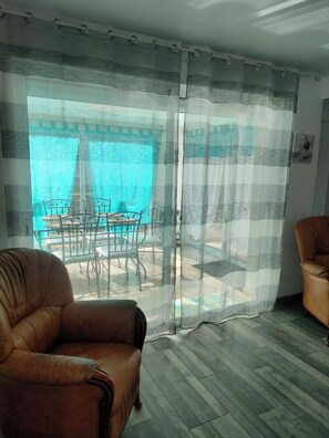 Ein Zimmer mit einem braunen Ledersessel, Holzboden, einer Glastür zu einem Außenbereich mit Tisch und Stühlen und Blick auf ein blaues Gewässer.