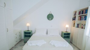 Quarto bem arrumado com uma cama grande, duas mesas de cabeceira com abajures, uma mesinha lateral verde, uma estante e um guarda-roupa.