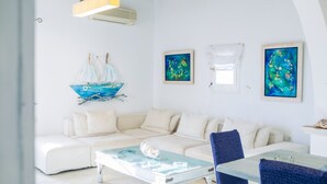 Uma sala de estar moderna com um sofá branco, uma mesa de centro de vidro e uma poltrona azul. Há quadros nas paredes e uma decoração náutica.