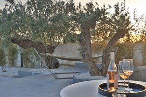 An outdoor seating area with a table, a bottle, and two glasses of wine, surrounded by olive trees and a hammock.
