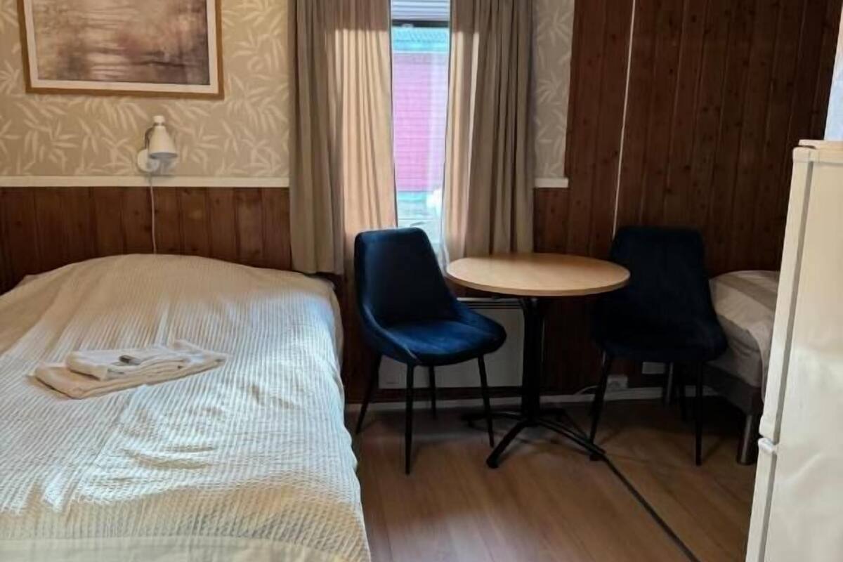 Habitació amb un llit, dues cadires, una taula petita, una làmpada i una finestra amb cortines.
