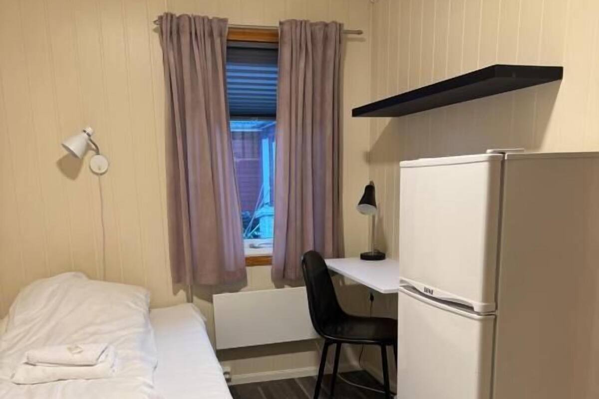 Habitació senzilla amb llit, escriptori, cadira, nevera i finestra amb cortines.