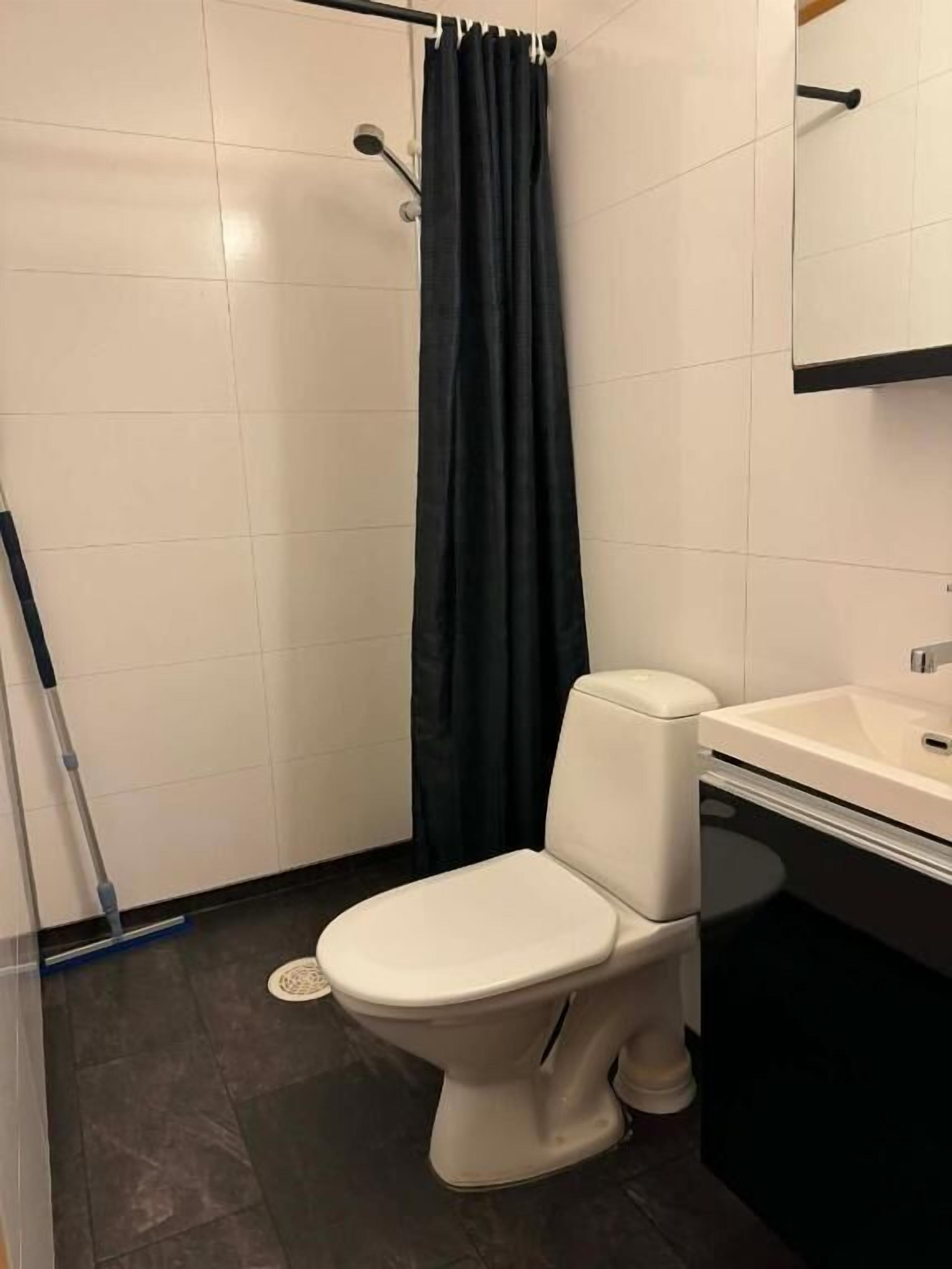 Un baño con inodoro, cortina de ducha, escoba y lavabo.