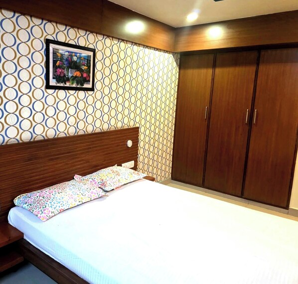 A bedroom with a wooden headboard, a bed with floral bedding, a wardrobe, and a patterned wall. - Lotus Park Inn (Bengaluru)