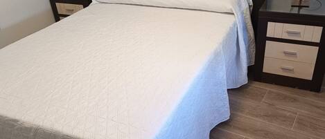A single bed with a white quilt, a wooden headboard, two bedside tables with lamps, and a wooden floor.
