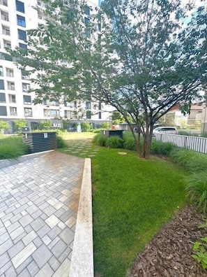 A paved area with a tree and grass, surrounded by a fence and a building in the background. - Soulful Apartment Mara M11 (Timișoara)