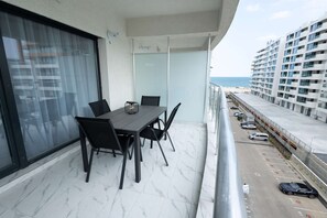 Een balkon met een eettafel en stoelen, uitzicht op het strand, en een parkeerplaats met auto's.