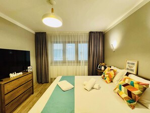 Quarto de hotel com cama, televisão, mesa de cabeceira, abajur e janela com cortinas.