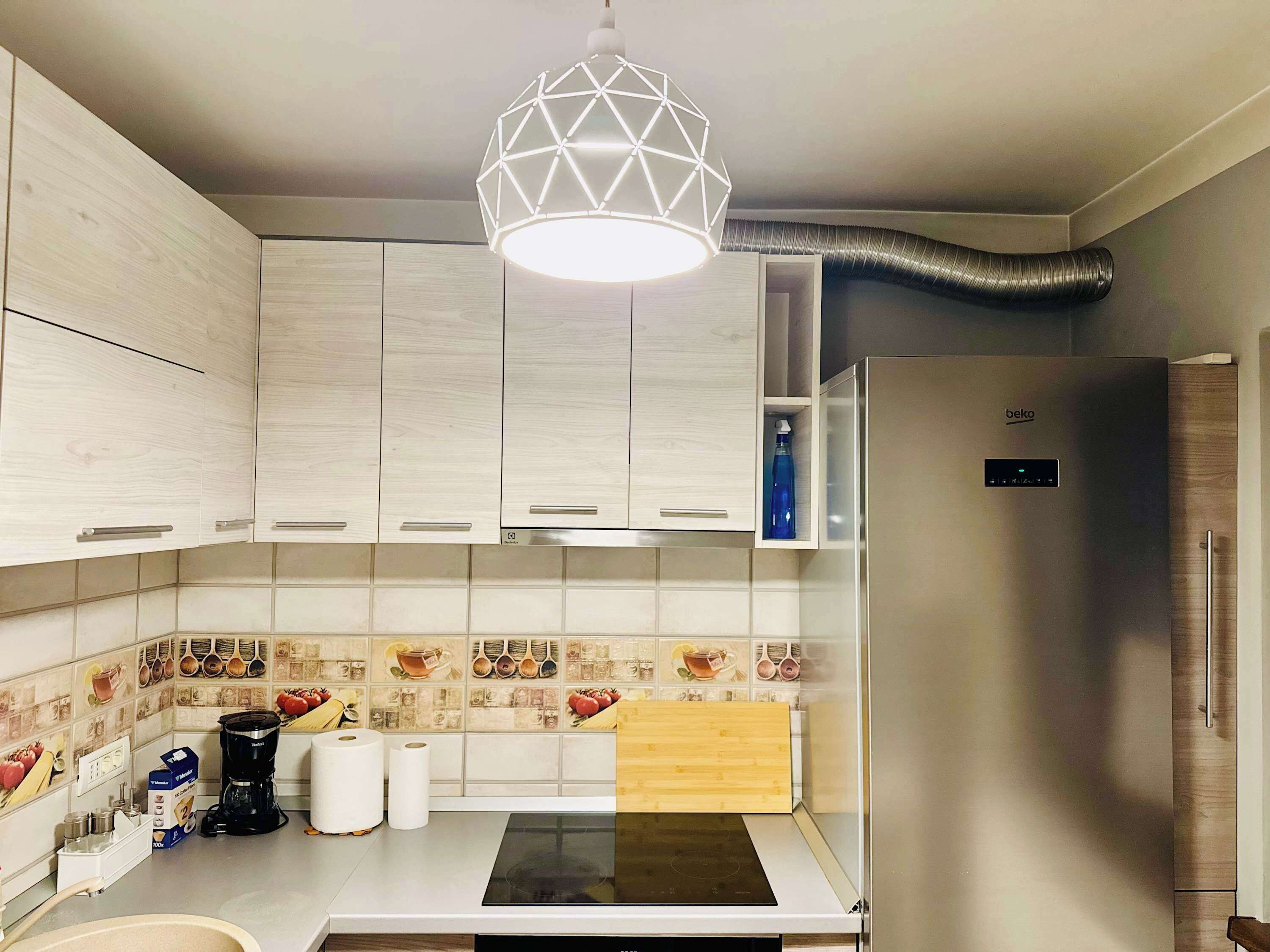 Una cocina moderna con armarios de madera clara, estufa integrada y una original lámpara colgante geométrica.