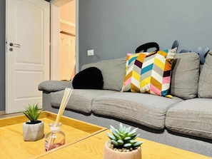 Sala de estar com um sofá cinzento, uma almofada colorida em padrão geométrico, uma pequena planta em vaso e uma garrafa de vidro com canudo.