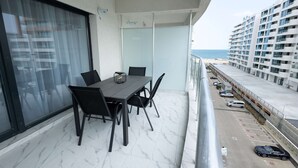 Un balcón con mesa y sillas, vista a la playa y un estacionamiento con coches.