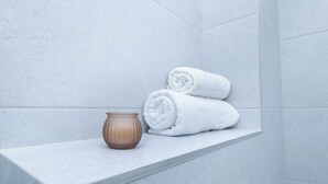 Un baño con baldosas blancas, un estante con toallas enrolladas y un jarrón marrón.