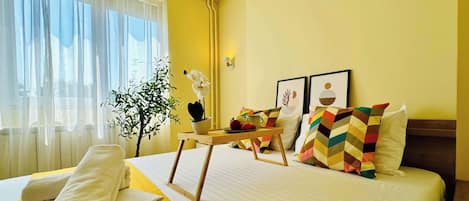 A hotel room with a bed, a table with a tray, a potted plant, and a window with curtains.
