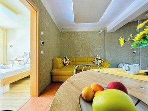 Ein modernes Interieur mit einer Couch, einem Esstisch mit Früchten und Blick auf einen Raum mit Bett und Fenster.