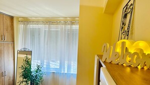 A room with a wooden cabinet, a mirror, a potted plant, a window with sheer curtains, and a 'Welcome' sign on the counter. - Joy City Stay Ultracentral Charming (Timișoara)