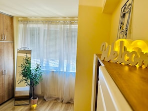 A room with a wooden cabinet, a mirror, a potted plant, a window with sheer curtains, and a 'Welcome' sign on the counter. - Joy City Stay Ultracentral Charming (Timișoara)