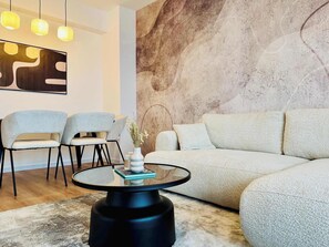 Un salon moderne avec une table basse ronde en verre, deux fauteuils gris, un canapé beige et un mur à texture.