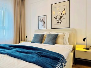 A hotel room with a bed, two pillows, a blue blanket, a nightstand with a lamp, and two framed artworks on the wall. - Joy City Stay M11-81 (Timișoara)