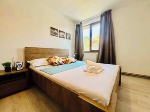 A bedroom with a wooden headboard, a bed with white bedding, a blue throw, and a rolled towel. There are two wall-mounted pictures, a nightstand with a plant, and a window with curtains. - Joy City Stay Victor 14 (Timișoara)