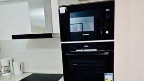 Une cuisine compacte équipée d’un four à micro-ondes et d’un four Bosch, d’une hotte en acier inoxydable et d’un plan de travail blanc avec une machine à café.