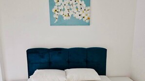 Un lit bien fait, avec des draps blancs et deux serviettes enroulées, une tête de lit rembourrée de couleur bleu foncé et un tableau représentant des fleurs blanches accroché au mur.