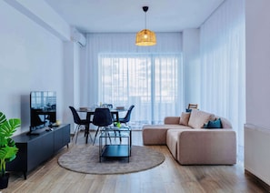 A modern living room with a dining area, a sofa, a TV, and a large window with sheer curtains.