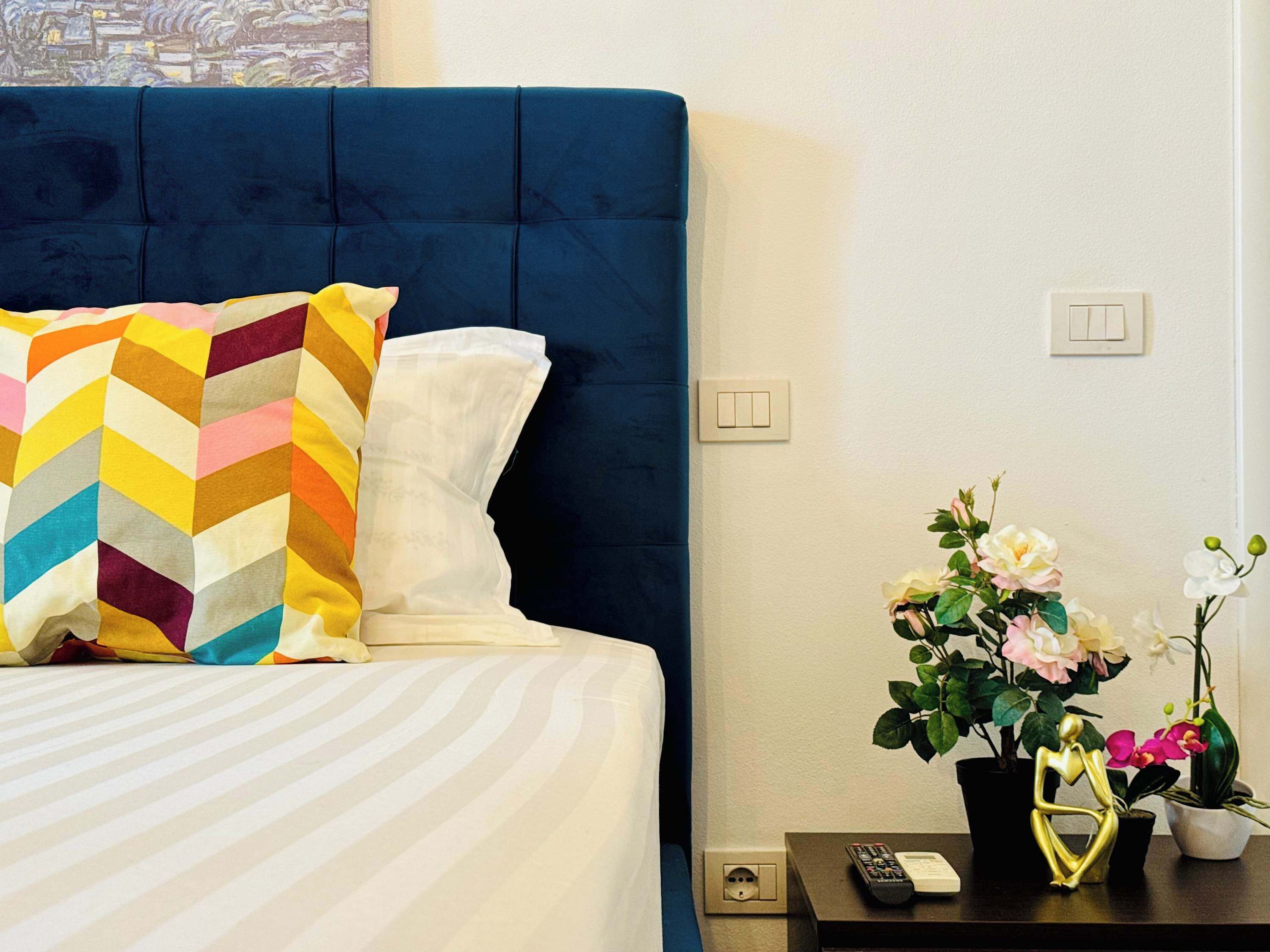 Una cama con cabecera azul, un cojín a cuadros de colores y un cojín blanco. Hay una mesita de noche con un control remoto, un teléfono y una planta en maceta con flores.